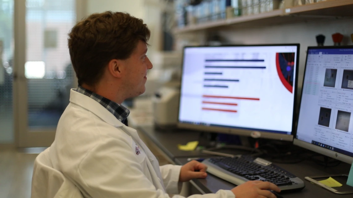 A man in a white lab coat looking at a pair of computer monitors.